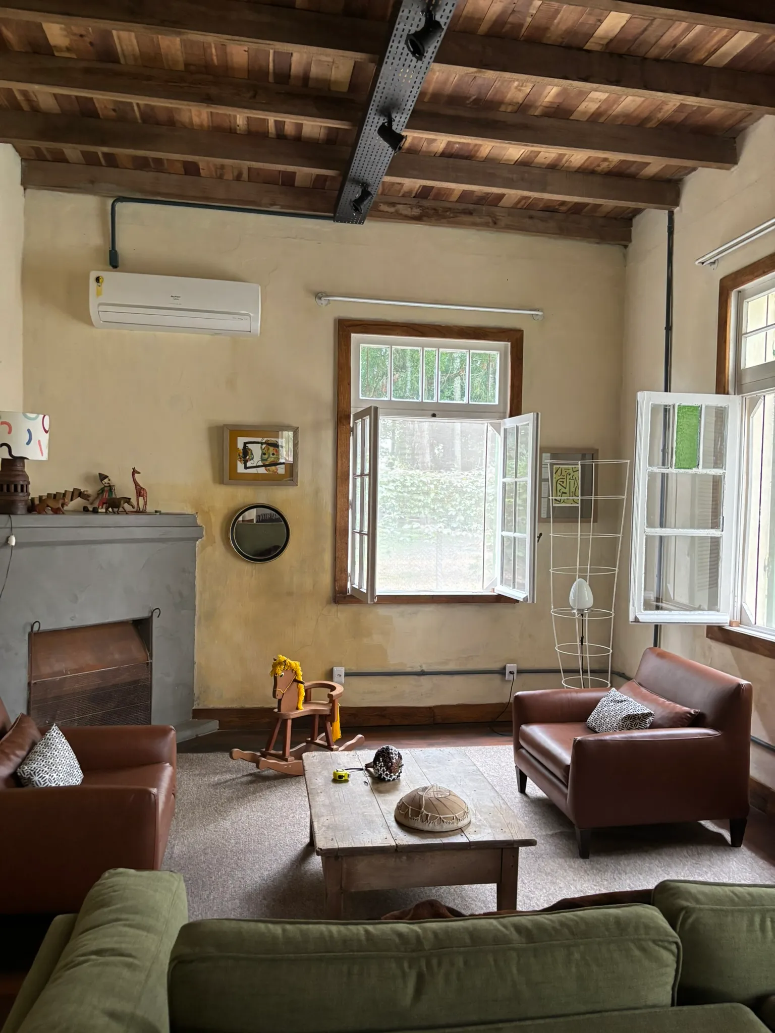 Sala de estar da Casa Micael: poltronas de couro, lareira, teto de vigas de madeira e janelas abertas para o jardim.